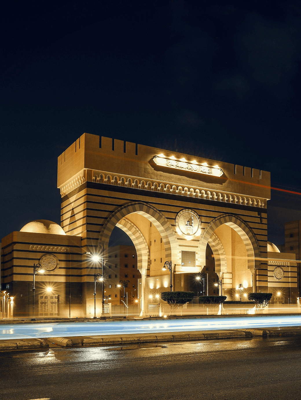 Grand illuminated stone archway with Islamic architectural details at night, warm golden lighting and light trails from traffic in the foreground